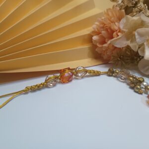 Strap para celular trabalhado em macramê com fio de seda na cor dourada com cristais dourados e incolores, contas esféricas na cor marrom translúcido e contas cúbicas na cor coral, contas métalicas na cor ouro velho